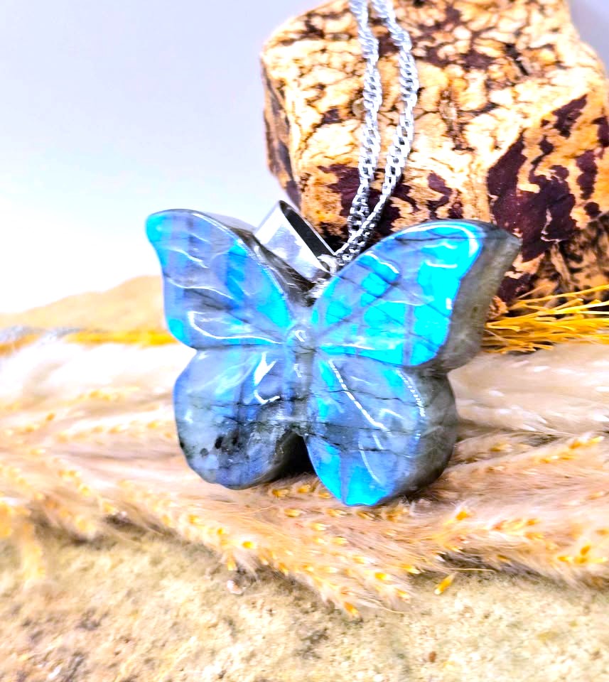 A butterfly-shaped pendant necklace made from polished blue labradorite stone with a silver chain, displayed on a textured cork block and beige fur against a soft-focus background.