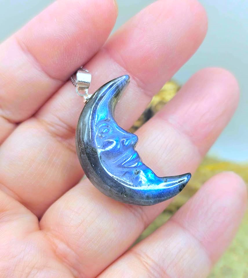 A crescent moon pendant with a smiling face, featuring a blue and purple iridescent labradorite finish, held in a person's hand.