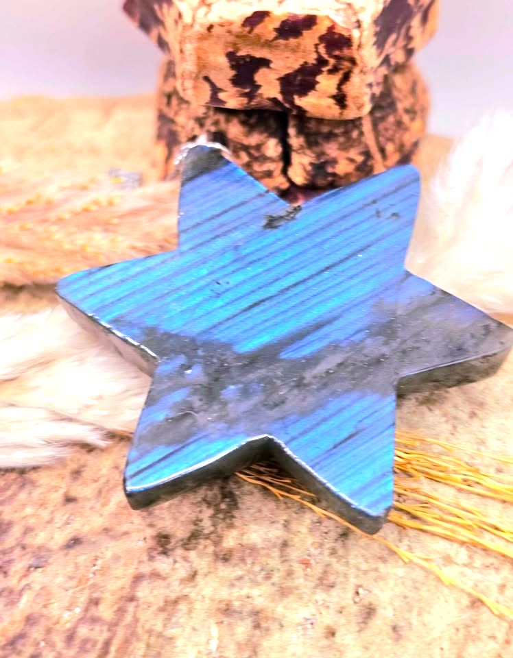 A star-shaped polished stone pendant with vibrant blue and green iridescent coloring, resting on a textured stone surface with decorative fur and a small wooden block in the background.