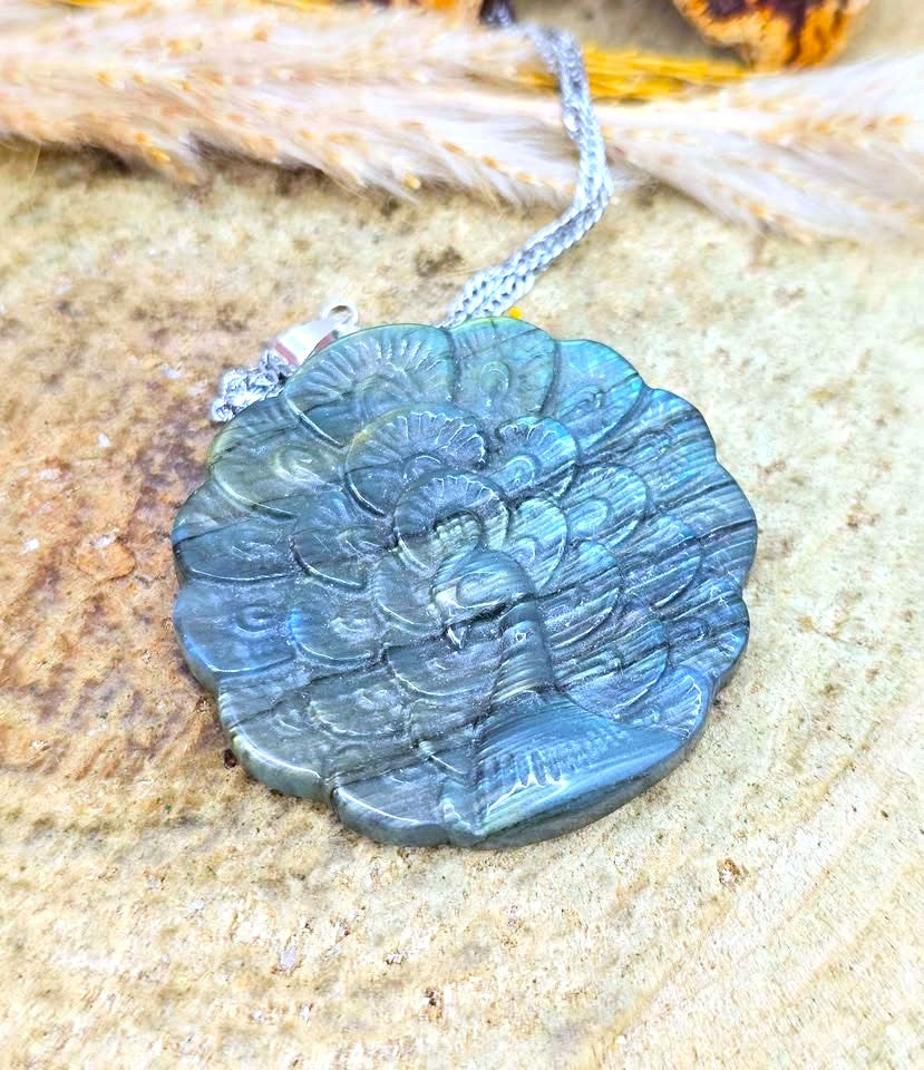 A round labradorite pendant necklace with a carved floral design, resting on a textured stone surface with dried grass in the background.