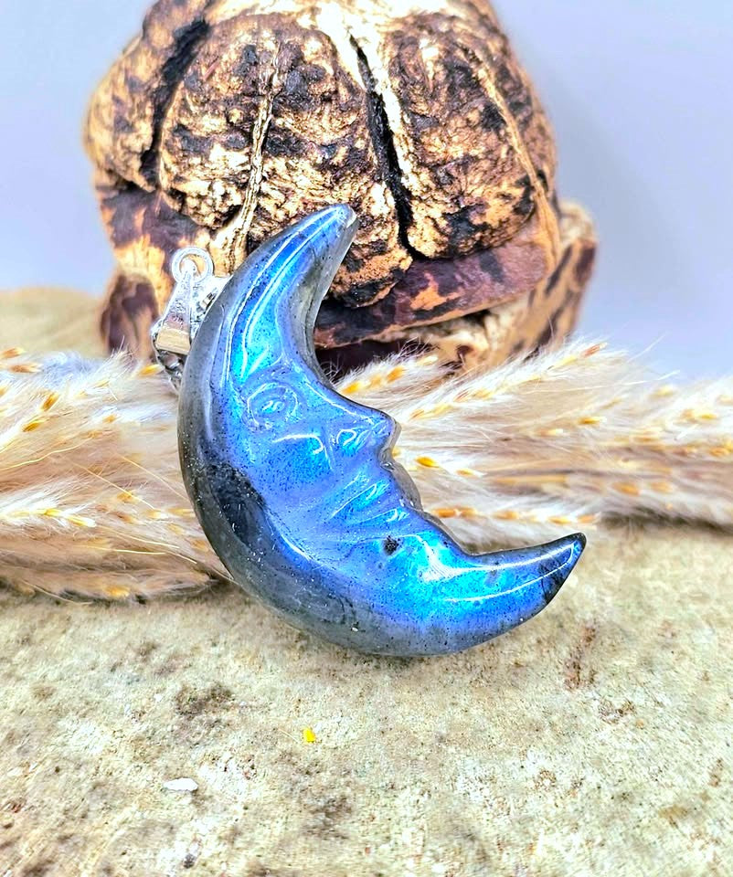 A crescent moon pendant with a vibrant blue iridescent finish and a small silver bail, resting on dried wheat stalks in front of a textured brown decorative object.