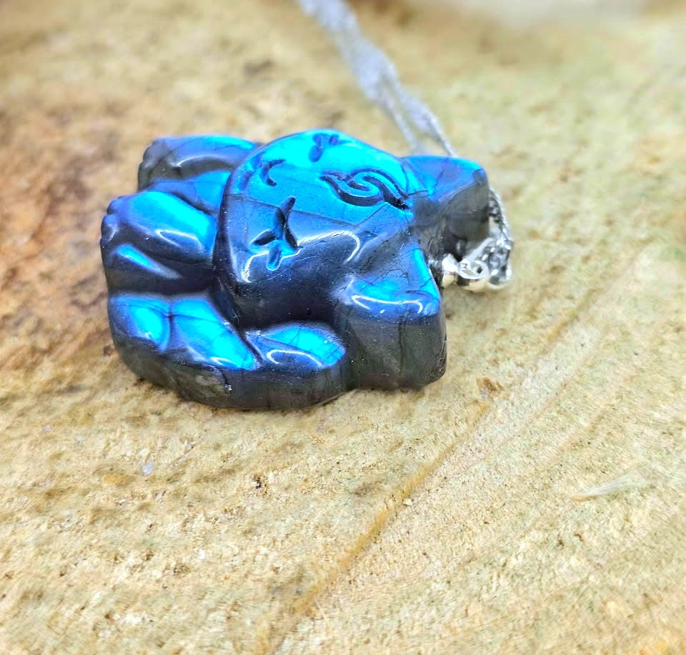A close-up of a small carved pendant or charm with a bright blue and black marbled pattern, attached to a silver chain, resting on a sandy beige surface.
