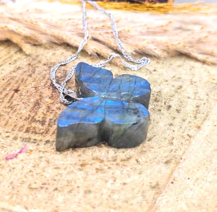 A silver chain necklace with a four-leaf clover pendant made of polished labradorite stone, resting on a textured sandy surface with dried leaves in the background.