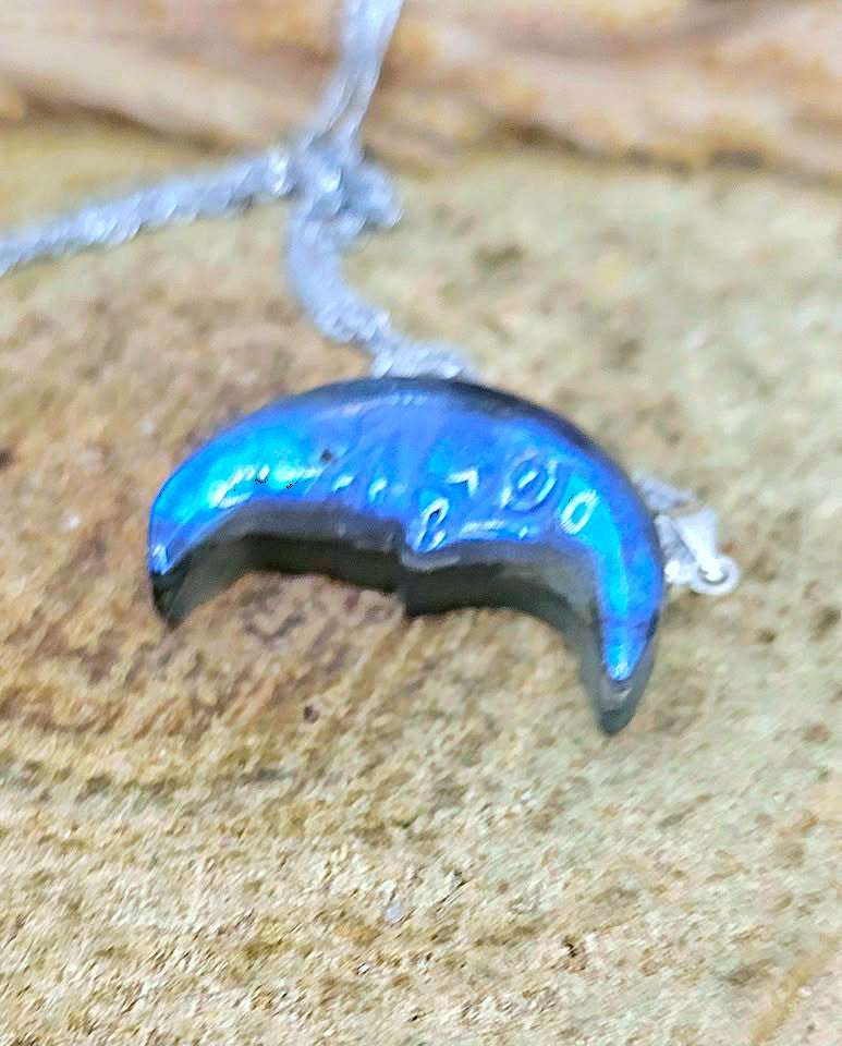 A silver crescent moon pendant with a vibrant blue iridescent opal inlay, resting on a textured sandy surface.