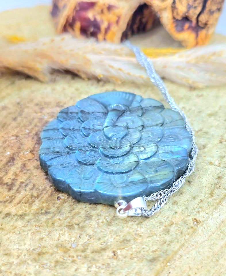 A round, textured pendant necklace made of layered blue-green stone with a silver chain, displayed on a sandy surface with dried grass and a blurred natural background.