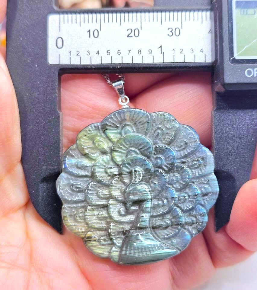 A close-up of a hand holding a carved oval pendant made of iridescent labradorite, positioned under a digital caliper to show its dimensions. The pendant features a detailed peacock design with shimmering blue, green, and gold tones.