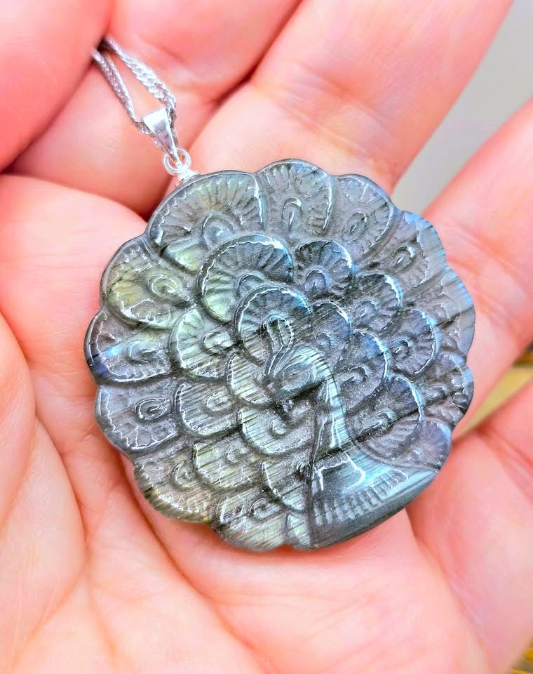 A close-up of a hand holding a round silver pendant necklace featuring a detailed carved peacock design. The pendant is made of a metallic material with a cool gray and silver color, and is suspended from a silver chain.