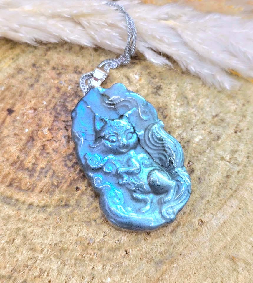 A silver pendant necklace featuring a hand-carved blue cat figurine, resting on a textured stone surface with white dried grass in the background.