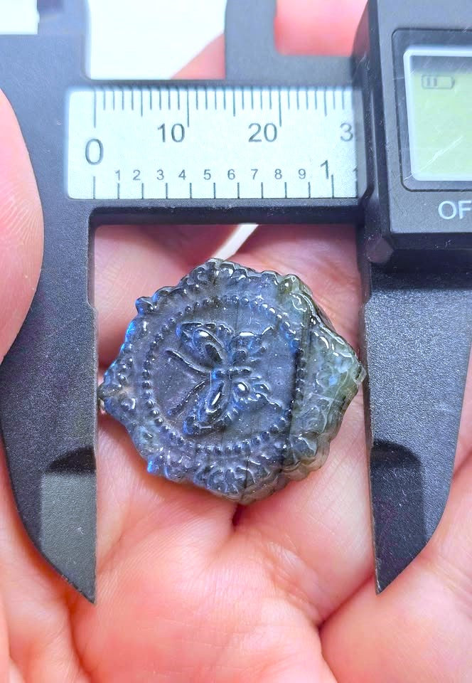 A close-up of a hand holding a small, antique-style embossed seal or medallion under a digital caliper to show its size. The medallion has a dark, weathered gray-brown appearance with a raised cross and floral motif.
