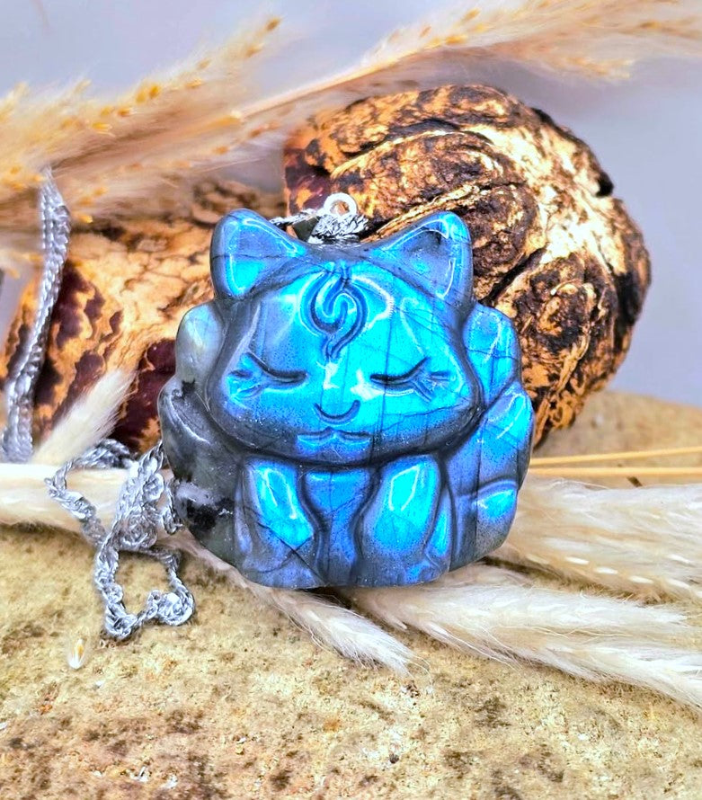 A blue cat-shaped pendant necklace with a silver chain, displayed on a textured surface with dried grass and a dried root.