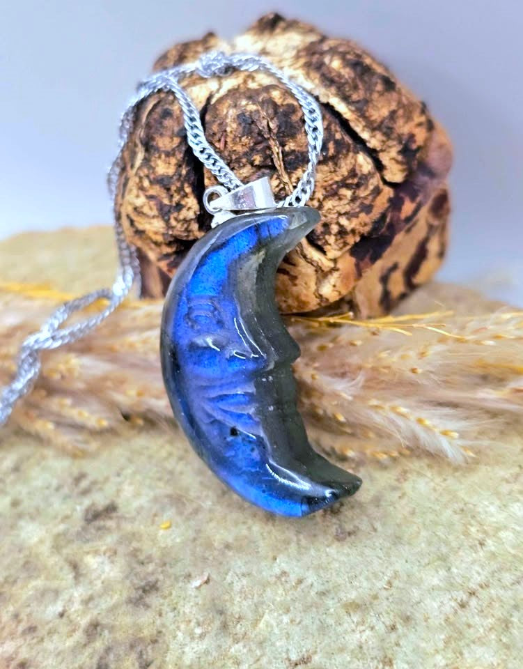 A crescent moon pendant necklace with a deep blue and black marbled pattern, suspended from a silver chain and styled on a textured rock with dried grass.