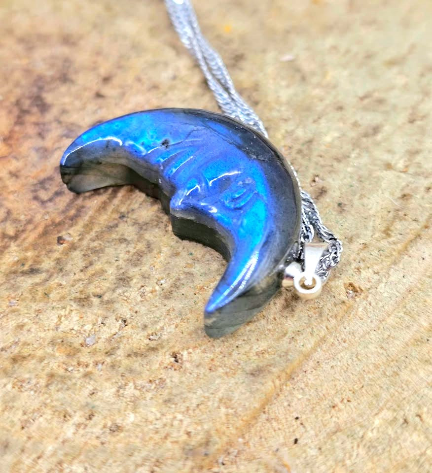 A crescent moon pendant necklace made of iridescent labradorite with a silver chain, resting on a textured brown surface.