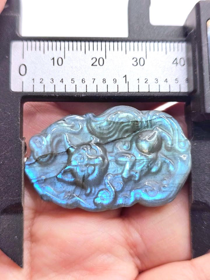 A close-up of a carved labradorite stone pendant held in a hand under a caliper, showing its blue iridescent finish and detailed relief carving.