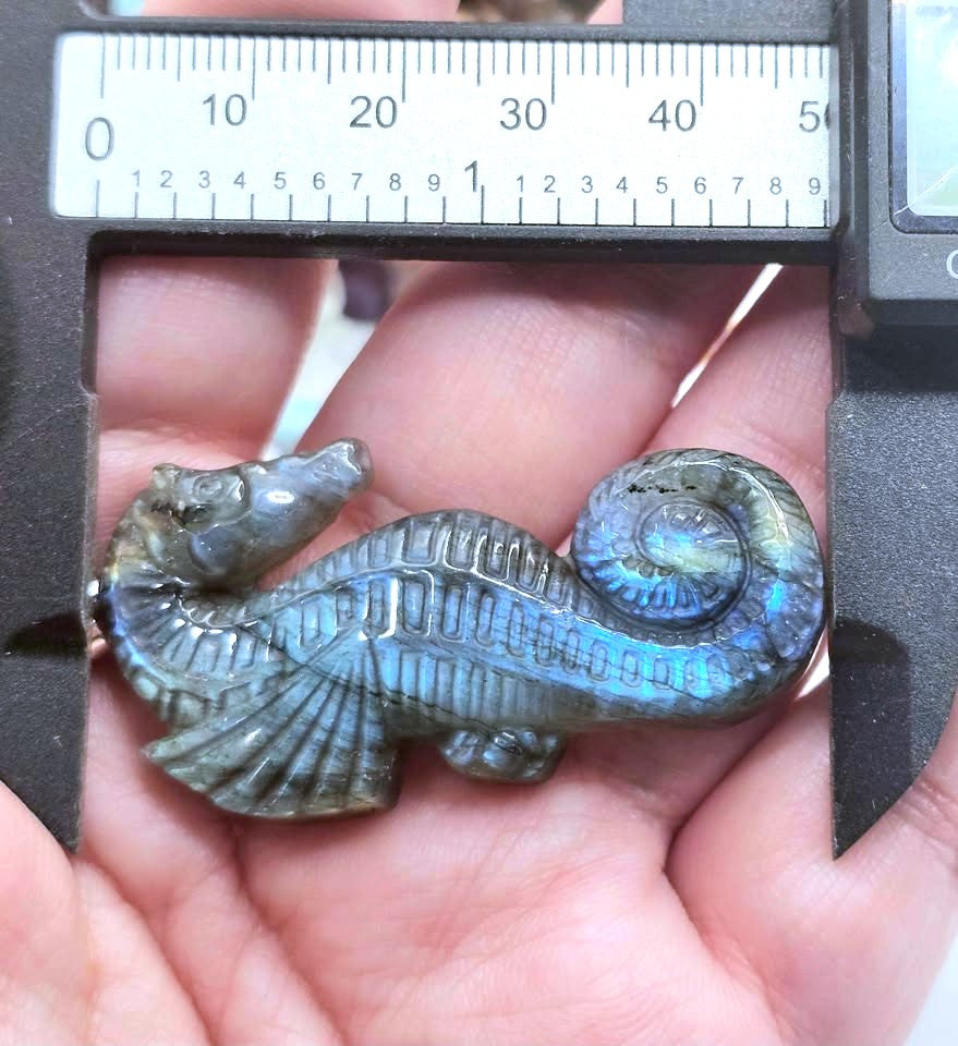 A seahorse-shaped polished stone pendant held in a person's hand under a caliper, showing an iridescent blue-green labradorite finish.