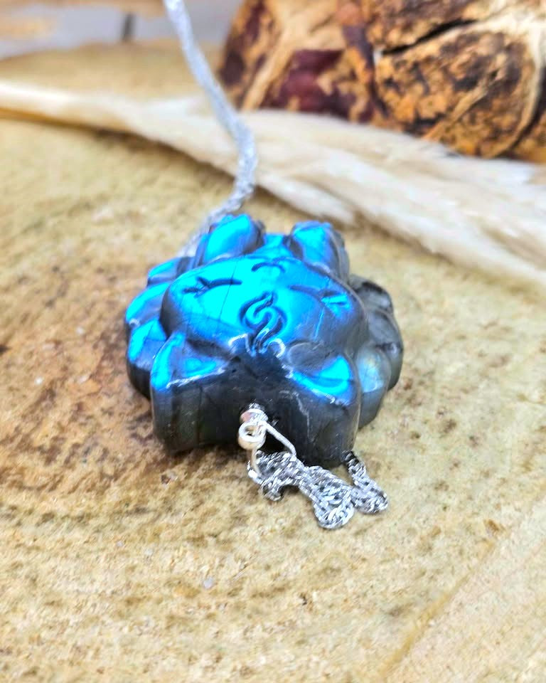 A close-up of a carved blue and black stone pendant with a silver chain and small silver dangling charm, resting on a textured tan surface.