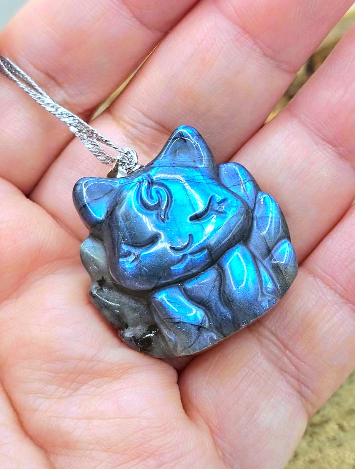 A hand holding a carved cat pendant made of iridescent labradorite stone, showing a blue and gray color shift, attached to a silver chain.