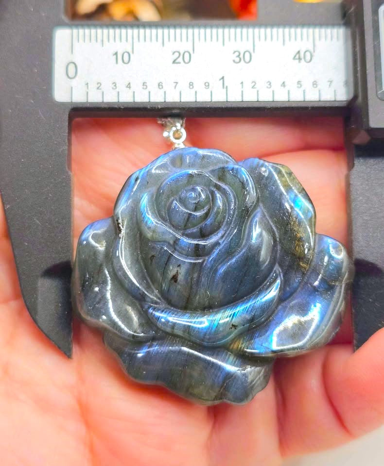 A polished labradorite rose-shaped pendant held in a hand under a caliper, showing its blue-gray color with iridescent flashes.