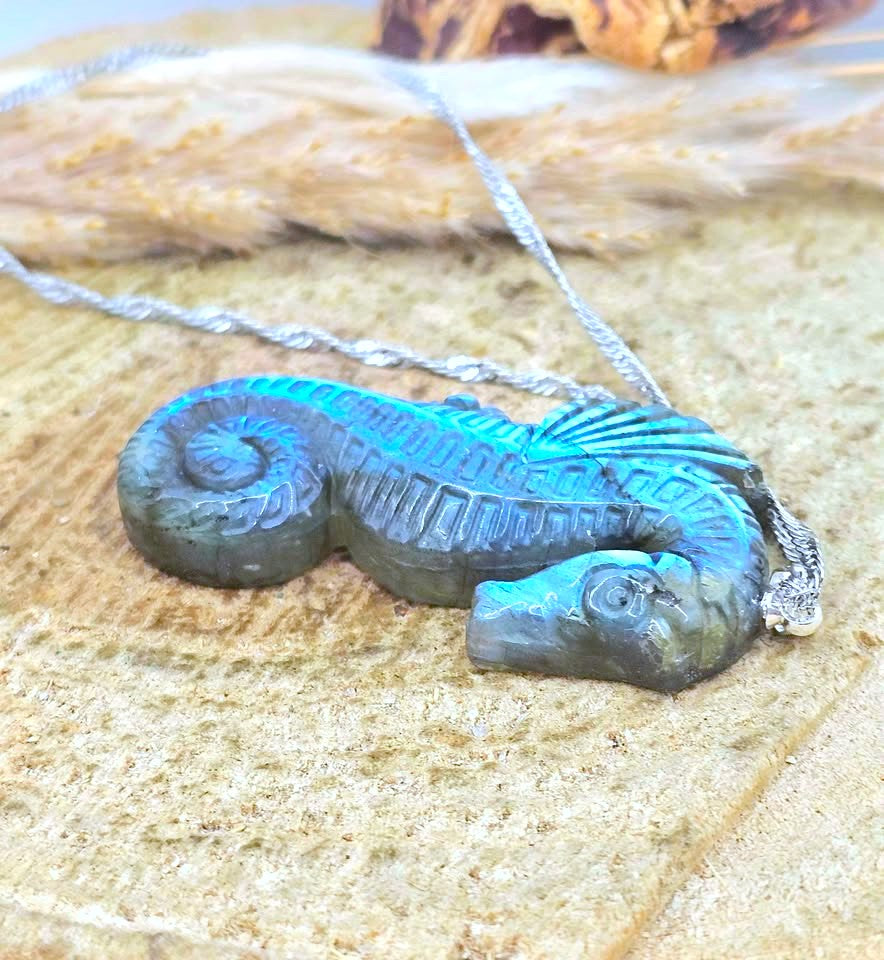 A seahorse-shaped pendant necklace with a textured, metallic teal and blue finish, resting on a sandy surface with a silver chain.