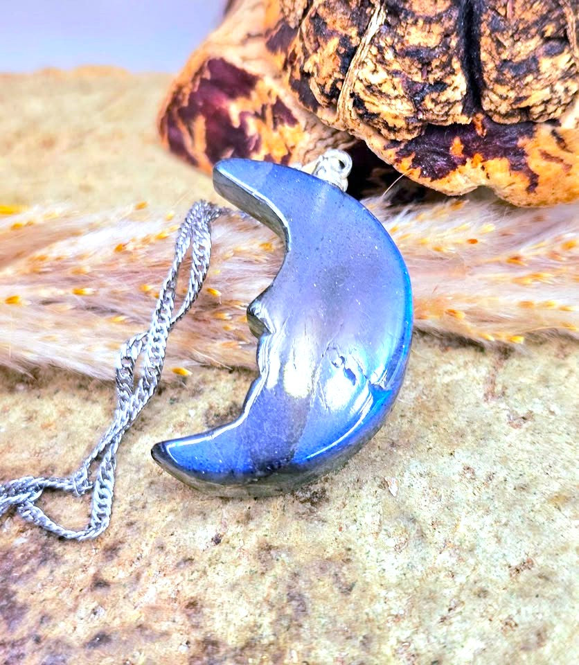 A crescent moon pendant necklace with a metallic blue and silver iridescent finish, resting on a textured stone surface next to a dried leaf and a silver chain.