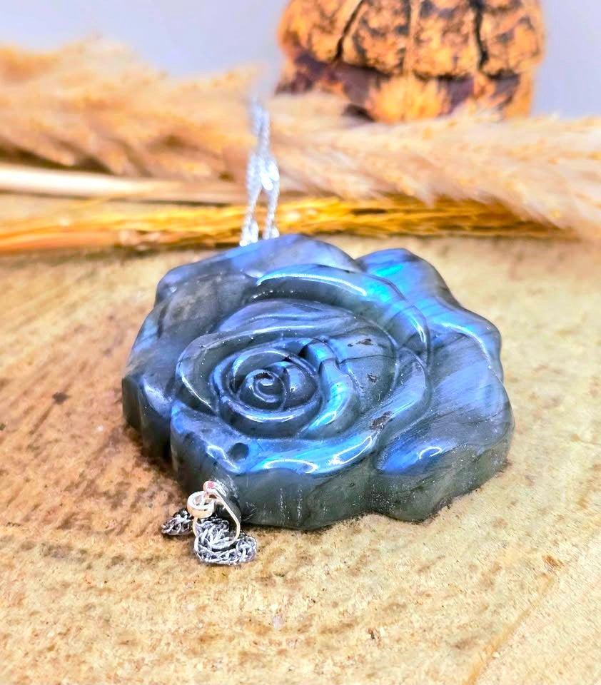 A carved labradorite rose pendant necklace with a silver-toned chain, displayed on a textured beige surface with dried grass and a small decorative object in the background.