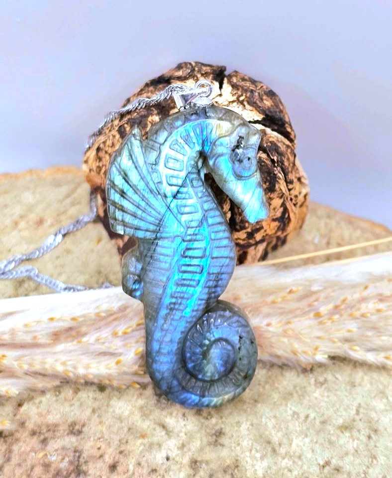 A seahorse-shaped pendant carved from iridescent blue labradorite, resting on a piece of driftwood with a silver chain attached, set against a natural background of wood and dried grass.
