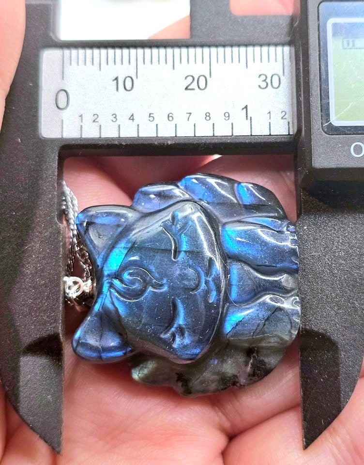 A close-up of a carved blue labradorite pendant held in a hand under a digital caliper to show its dimensions. The pendant features a stylized face with a silver-toned chain loop on the left side.