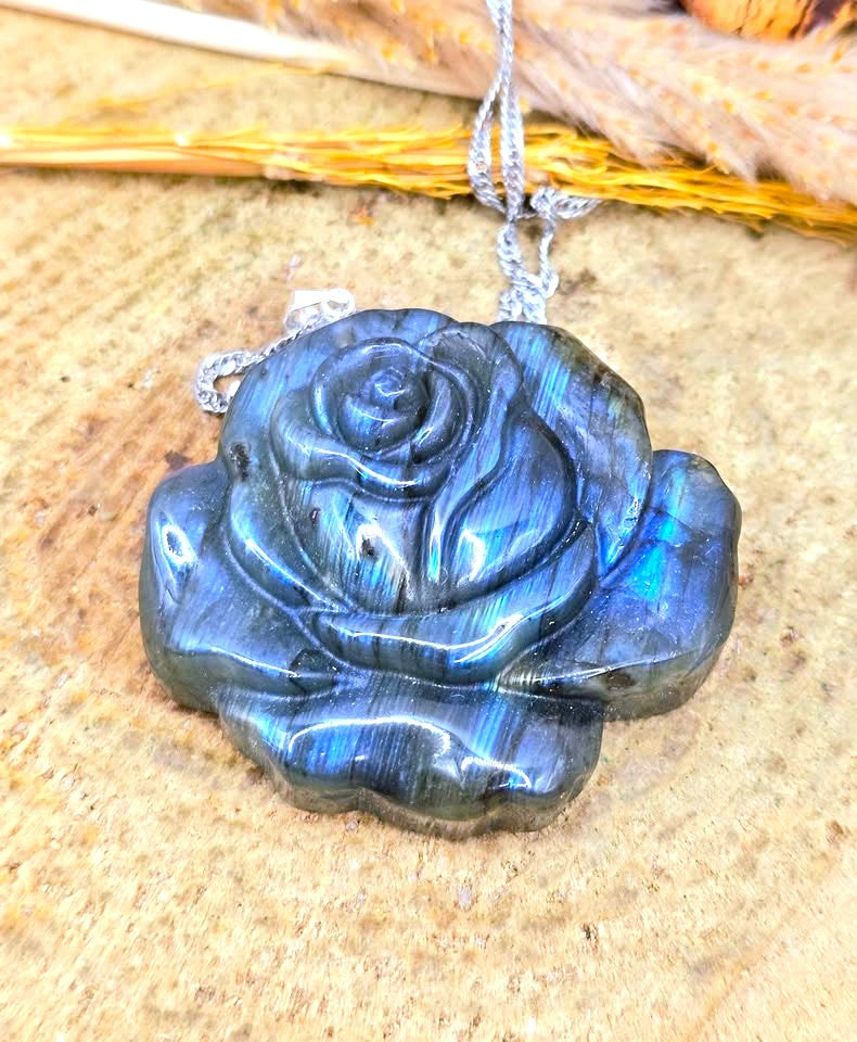 A carved labradorite rose pendant necklace with a silver chain, displayed on a textured wooden surface with dried grass.