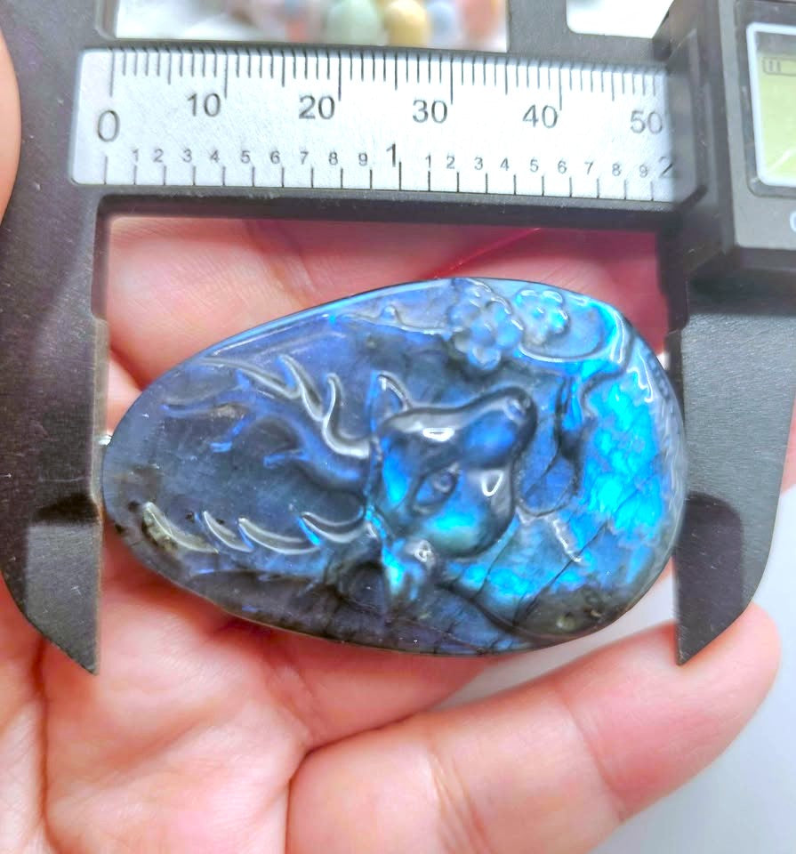 A teardrop-shaped labradorite stone pendant with a carved dragon design, held in a hand under a caliper to show its size. The stone displays a vibrant blue and green iridescent play-of-color.