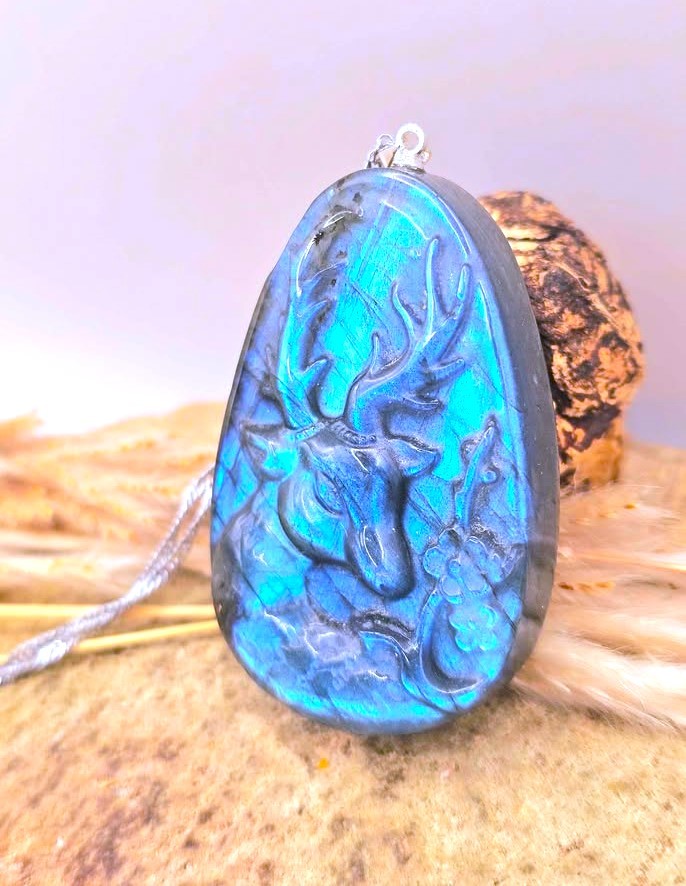 A teardrop-shaped carved pendant featuring a deer head design in vibrant blue and turquoise with dark gray shading, suspended from a silver chain and resting on a textured surface.