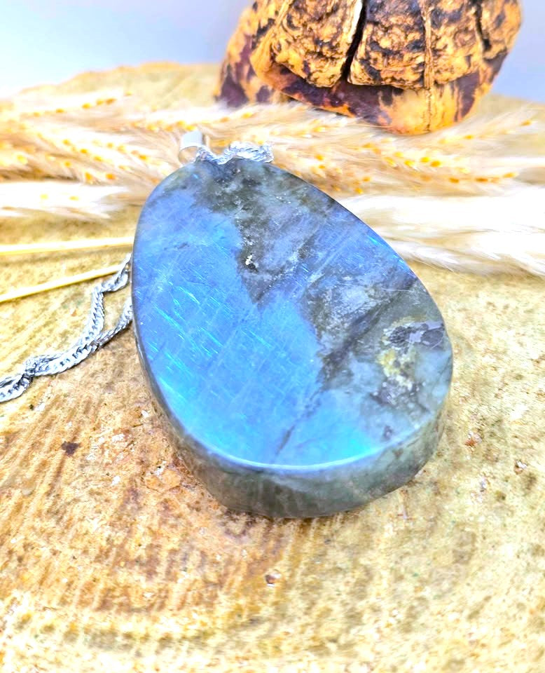 A teardrop-shaped labradorite stone pendant with a silver chain, resting on a textured wooden surface with dried wheat stalks and a bark piece in the background.