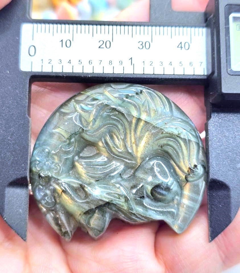 A close-up of a carved oval pendant or charm held in a hand, with a digital caliper above it showing its width. The object appears to be made of polished stone or resin with a swirling marbled pattern in gray, green, and gold tones.