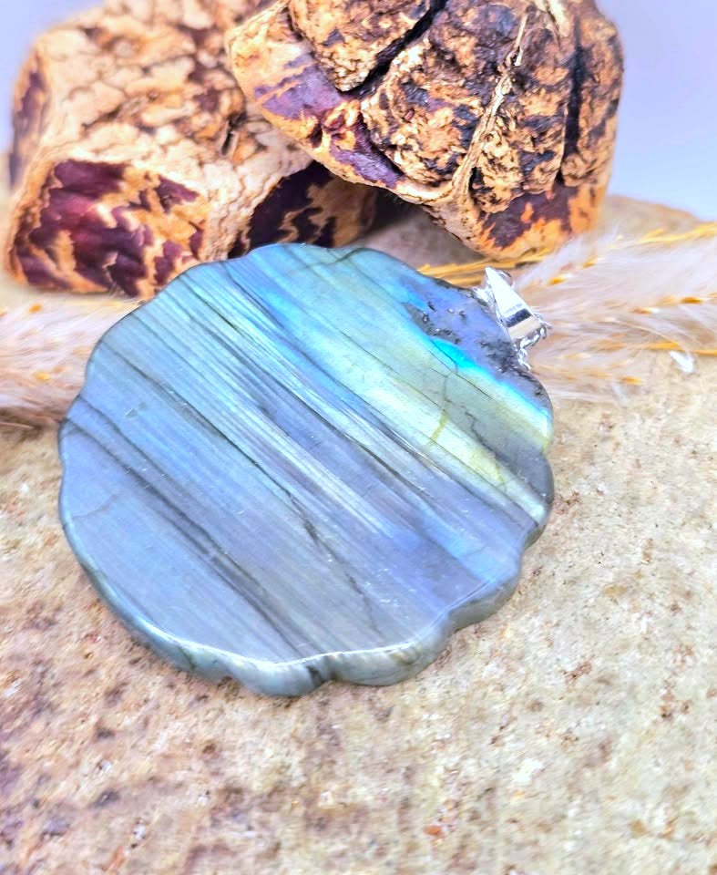 A scalloped oval labradorite pendant with a silver bail, resting on a textured stone surface with dried feathers and a piece of wood in the background.