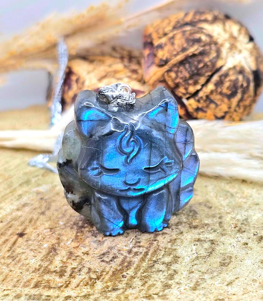 A carved cat-shaped pendant or charm made from iridescent labradorite, showing blue and green flashes, with a silver bail at the top, displayed on a textured wood surface with dried botanicals in the background.