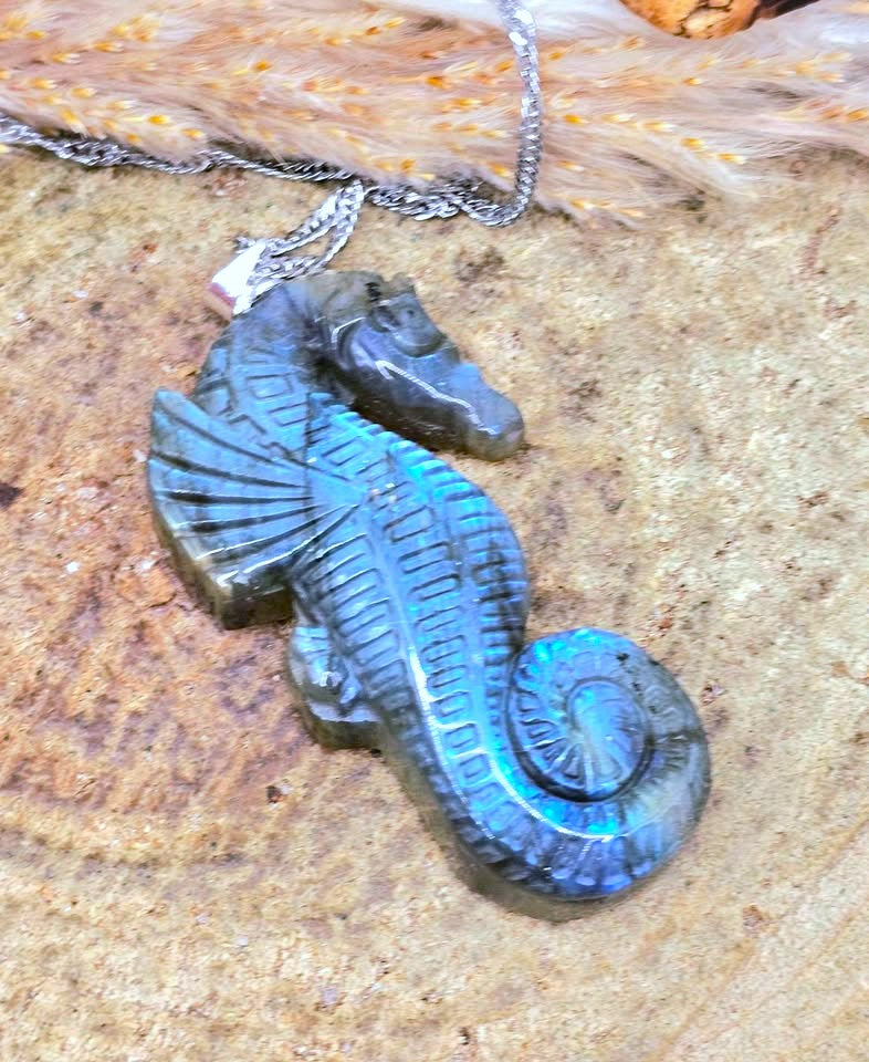 A carved seahorse pendant necklace made from iridescent blue-green labradorite stone, hanging from a silver chain on a textured sandy surface.