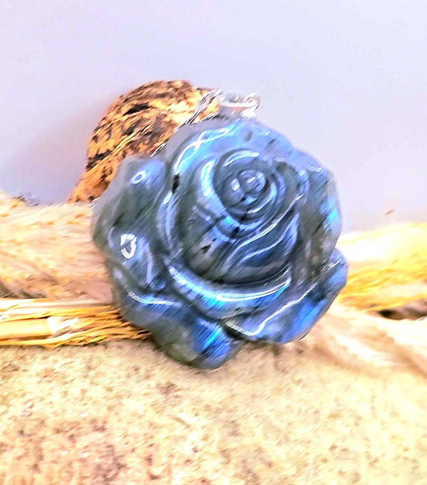 A carved blue labradorite rose pendant with a silver bail, displayed on a rustic background of dried grass and wood.