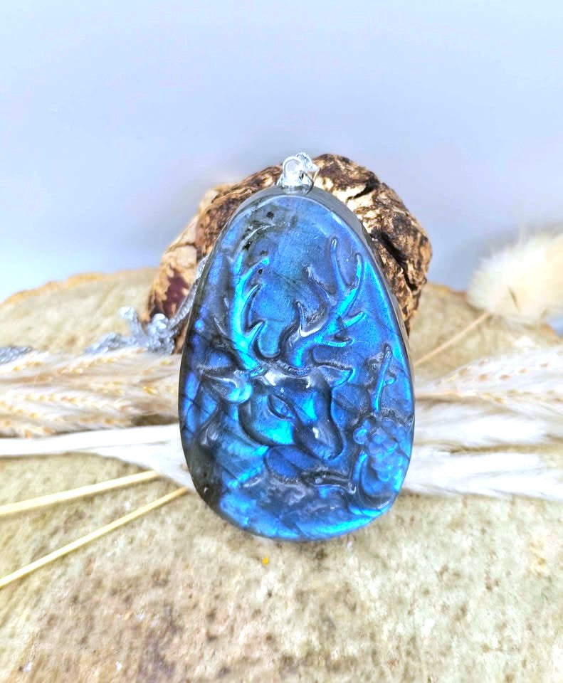 A teardrop-shaped labradorite pendant with a carved deer design, set in a rustic brown wooden ring, displayed on a stone surface with dried grass.