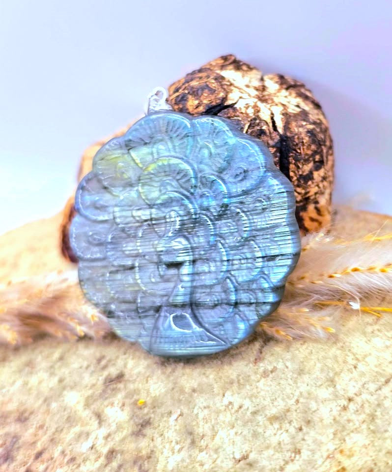 A round labradorite pendant with a carved peacock design, displaying a blue and green iridescent flash, resting on a piece of wood with feathers against a light background.