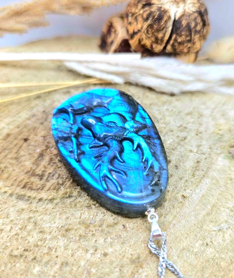 A polished blue labradorite pendant with an embossed animal design, hanging from a silver chain against a rustic wood background.