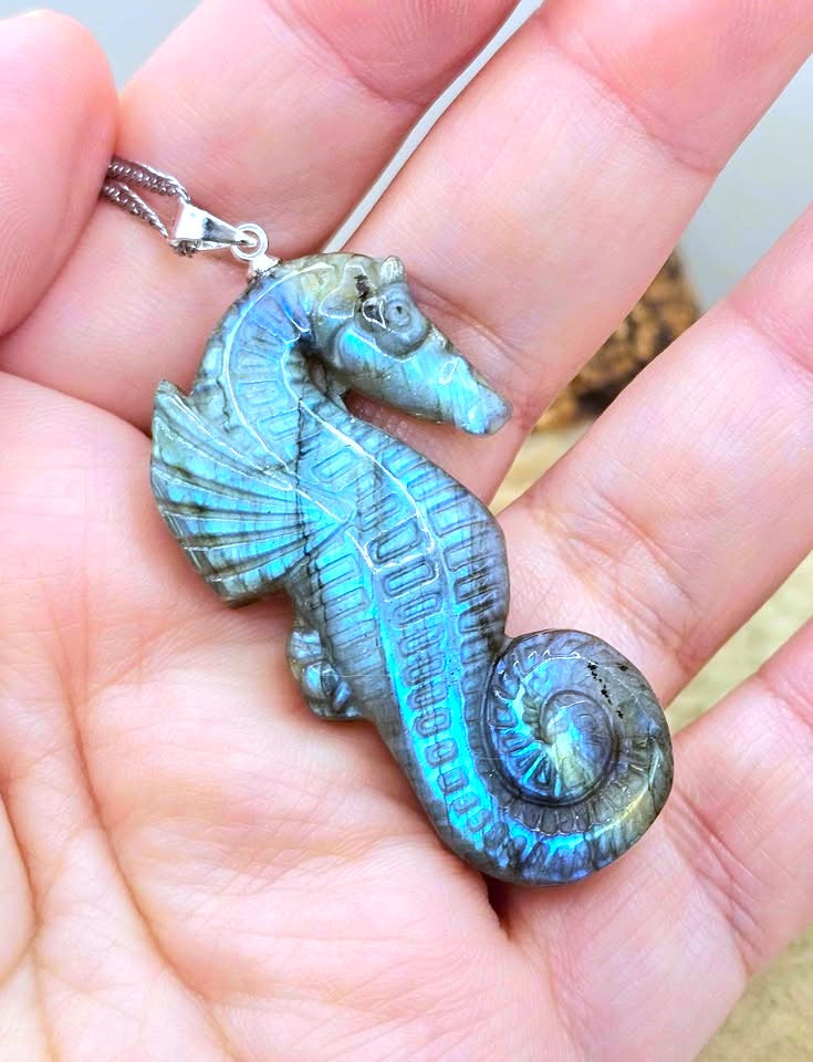 A seahorse-shaped pendant with a metallic teal and blue iridescent finish, held in a person's hand on a sandy background.