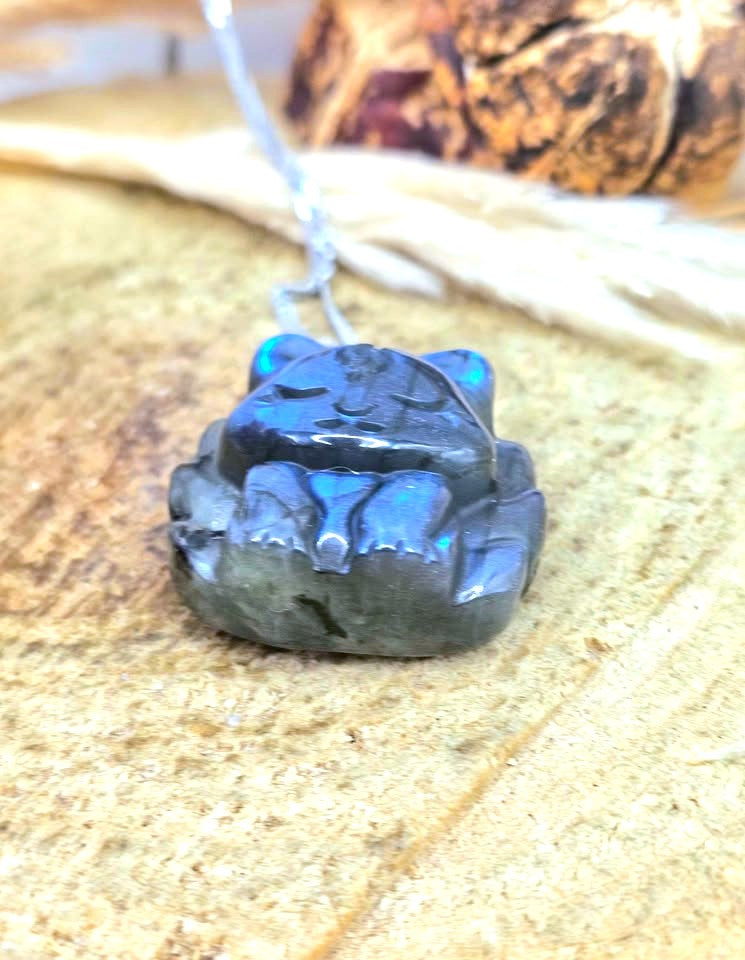 A small carved pendant or charm with a cat-like face and paws, made from dark polished stone with strong blue iridescent highlights, hanging from a thin silver cord on a textured tan surface.