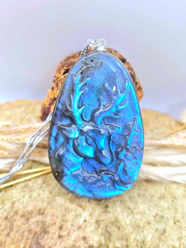 A teardrop-shaped pendant featuring a carved deer head design in vibrant blue and black iridescent stone, suspended from a silver chain and displayed on a textured stone surface.