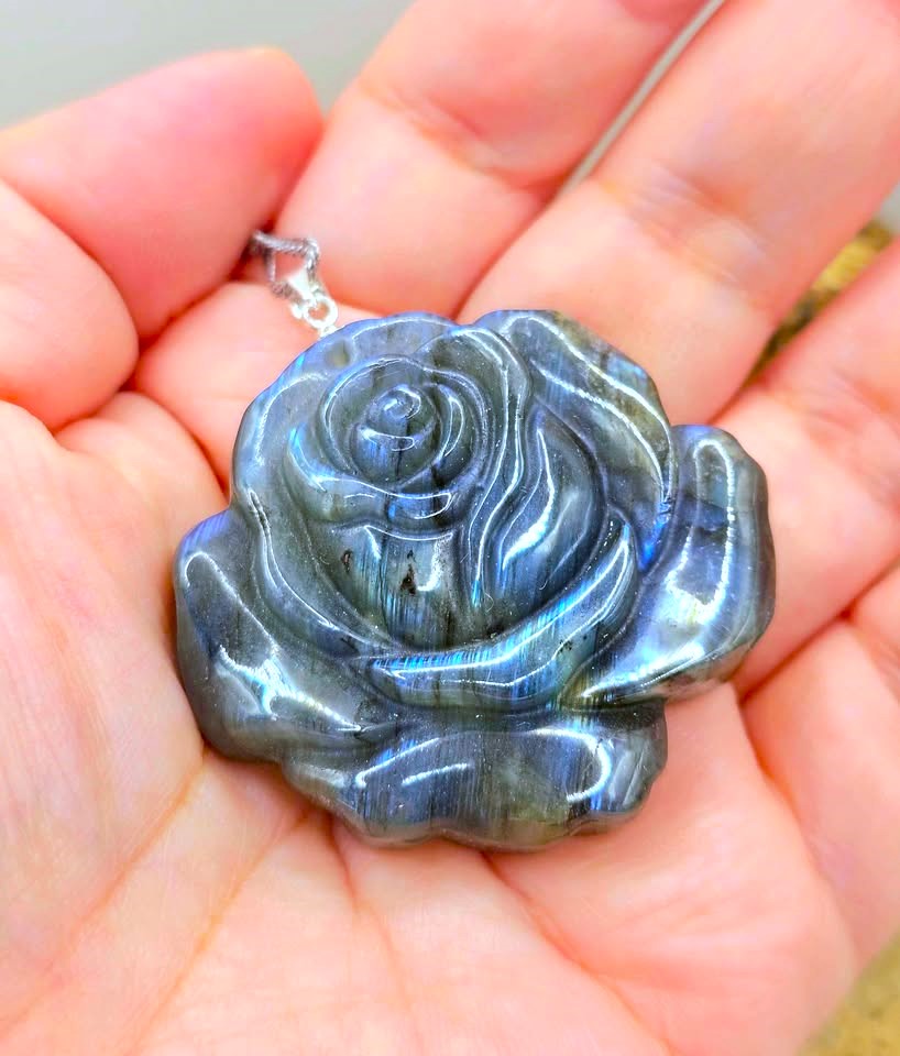 A carved labradorite rose pendant resting on a person's palm, showcasing its characteristic blue-gray iridescent shimmer.