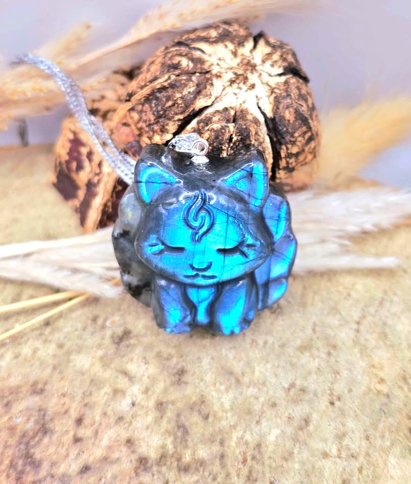 A carved blue cat pendant with a silver bail, resting on a textured stone surface with dried grass and a dried root in the background.