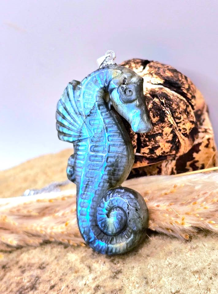 A seahorse-shaped pendant with a metallic blue and black textured finish, displayed in front of a brown seashell on a sandy surface.