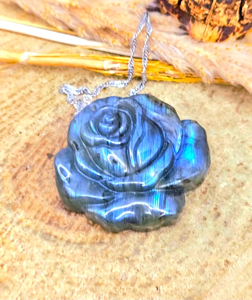 A carved blue labradorite rose pendant necklace with a silver chain, displayed on a textured wood surface with dried grass.
