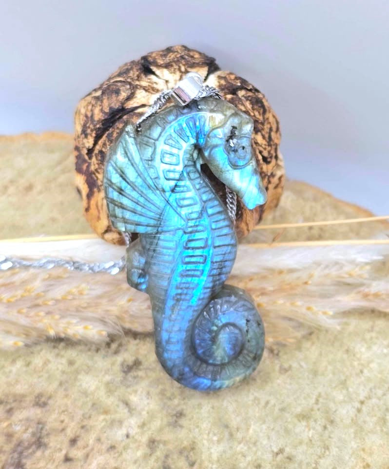 A seahorse-shaped pendant with a blue iridescent finish, attached to a silver chain and displayed against a natural seashell and sandy background.