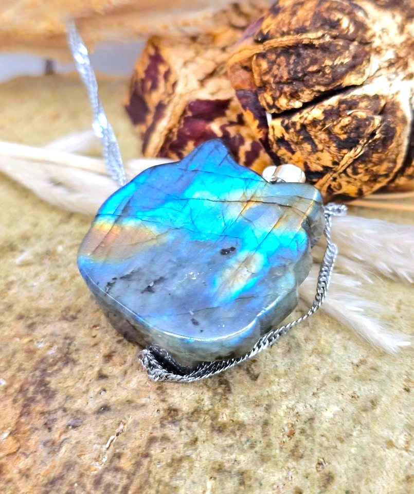 A raw labradorite gemstone pendant with vibrant blue, green, and orange iridescence, suspended on a silver chain, displayed on a textured stone surface with dried botanicals in the background.