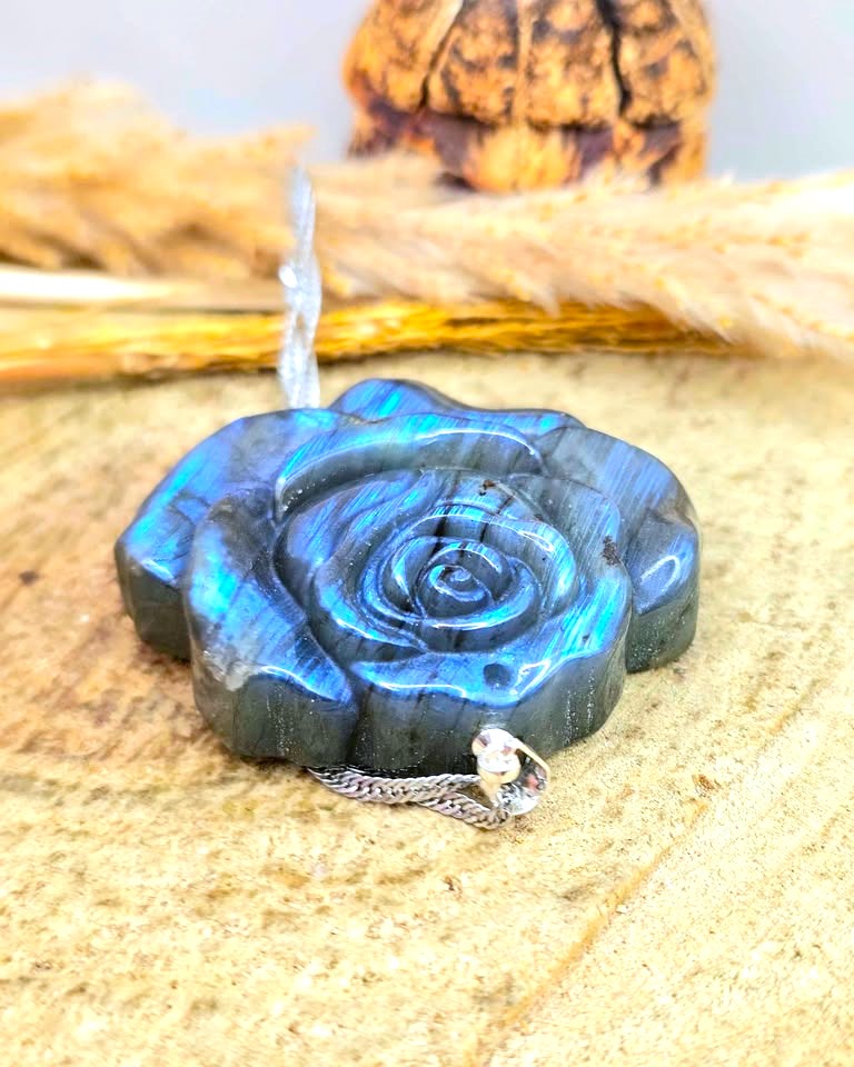 A carved labradorite rose-shaped pendant with a silver chain, displayed on a textured beige surface with dried grass and a pinecone in the background.