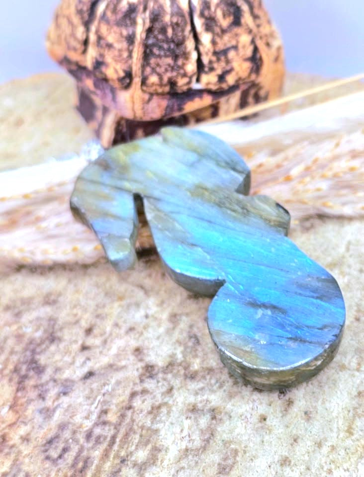 A polished labradorite stone pendant with a fish-shaped silhouette, displaying vibrant blue and green iridescent flashes, resting on a textured stone surface with a wooden object in the background.