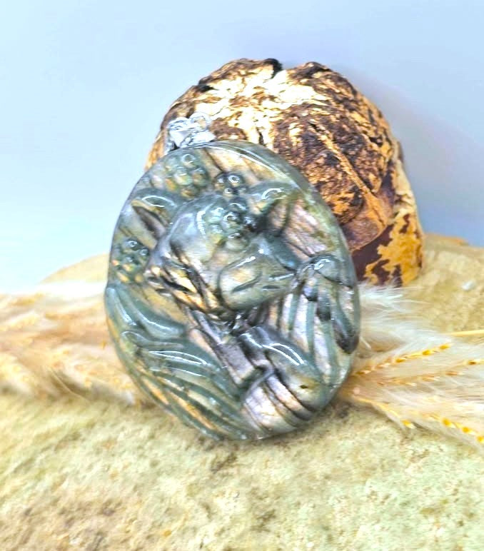 A round carved stone pendant or medallion featuring a detailed animal relief, displayed on a textured surface with a shell and dried grass behind it.